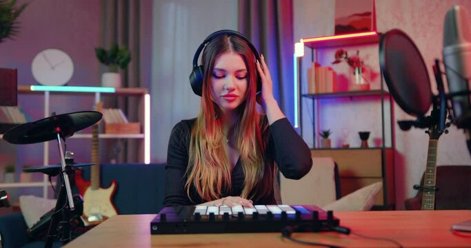 Modern music technology concept where lovely happy smiling professional female DJ artist in headphones creating new insrtumental music on drum pads midi player at home studio