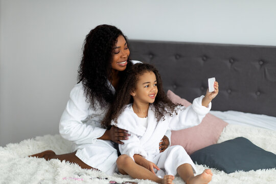 Cheerful black mother with daughter taking selfie in bath robes, posing for photo on bed at home