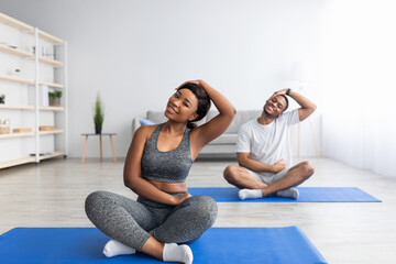 Sporty black couple exercising at home, sitting on sports mats, stretching their necks during domestic training