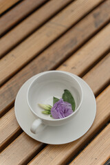 A white cup and saucer stand on a wooden background. There is a purple flower in the cup. Flower tea. Drink tea outside. Romantic mood.