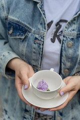 The girl is holding a white cup with a purple flower in her hands. Flower drink. Women's tea. Tea for weight loss. Women's tricks. A drink for a slim figure. Diet for a slim figure.