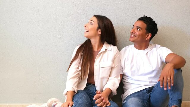 Young couple at home sitting on the floor looking up in to the air and smiling - concept of love and relationship wito mortgage and new life together - interracial boy and girl