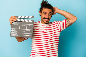 Young caucasian man with long hair holding clapperboard isolated on blue background touching back...