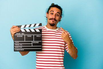 Young caucasian man with long hair holding clapperboard isolated on blue background pointing with...