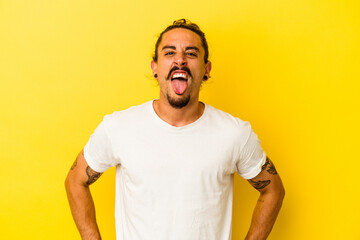 Young caucasian man with long hair isolated on yellow background funny and friendly sticking out tongue. - obrazy, fototapety, plakaty
