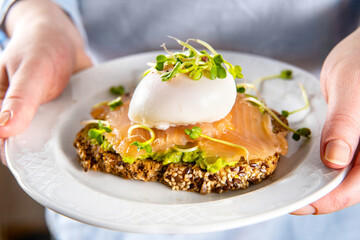 Unrecognisable woman in blue shirt holds a plate with salmon toast, avocado and posched egg. Close up. Healthy vegetarian breakfast food. Brain food concept.