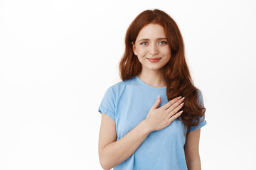 Fototapeta premium Portrait of natural redhead girl holds hand on heart and looks at camera happy, listening to national anthem, give her pledge, being touched or pleased, white background