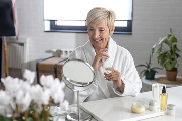 An older woman with short blonde hair in a white bathrobe sits at the table and puts cream on her...