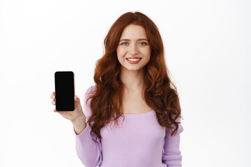 Fototapeta premium Smiling beautiful girl with red curly hair, showing application, demonstrate an empty smartphone screen and looking happy, standing against white background
