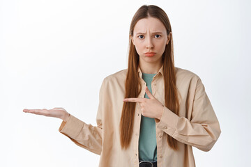 Disappointed frowning girl pointing at open hand, holding item and look with jealous or unfair...