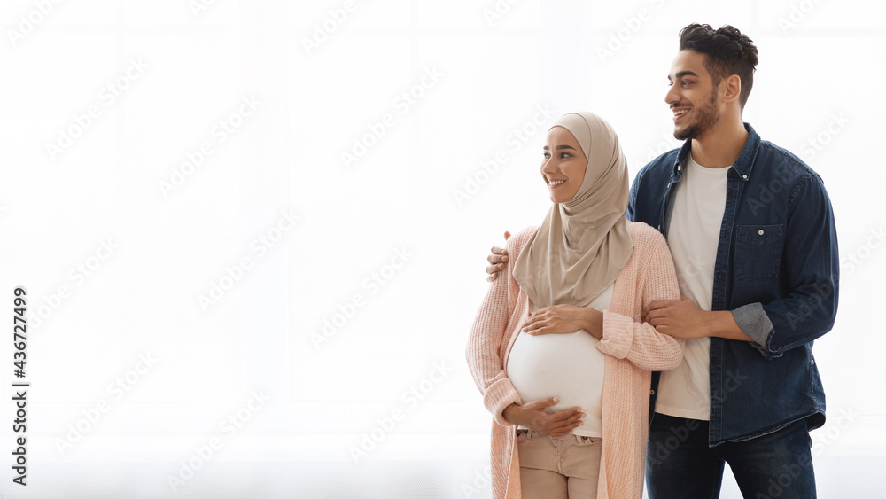 Wall mural young pregnant arabic family standing near window at home and looking aside