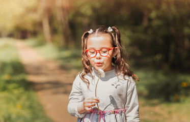 Little cute girl in glasses blowing on dandelion in nature in summer