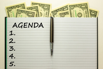 Agenda symbol. White note with the concept word 'agenda' on beautiful white table, metallic pen and dollar bills. Business, agenda concept.
