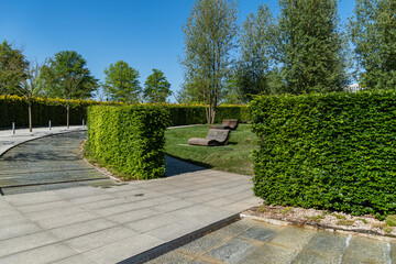 Fototapeta premium Artificial swift river with granite banks runs around evergreen hedge. Comfortable benches in shade of deciduous trees.Landscape city park of Krasnodar or 