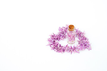 Glass bottle standing on a white background surrounded by lilac flower petals in the shape of a heart. Idea for floral fragrances.