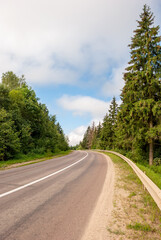 The road goes into the distance. Spruce forest and blue sky with clouds