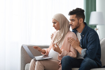 Portrait Of Confused Pregnant Muslim Couple Looking At Laptop Screen At Home