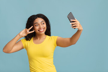 Excited african american lady taking selfie with smartphone and showing victory gesture on blue...