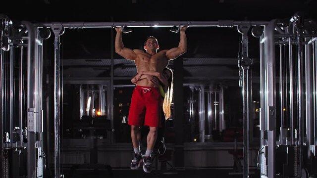 Man and woman training in the gym - a man trying to do pull ups with a woman hanging on his waist
