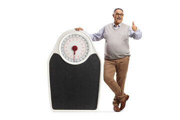 Full length portrait of a mature corpulent man leaning on a big scale and showing thumbs up