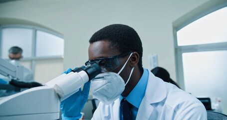 Close up of young African American busy male medical scientist working in laboratory on virus cure. Man specialist in mask and goggles looking at blood cells through microscope modern equipment