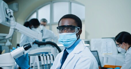 Close up of young serious African American man medical expert in mask sitting at desk in clinic lab working on modern microscope equipment and looking at camera. Medical scientist, covid treatment