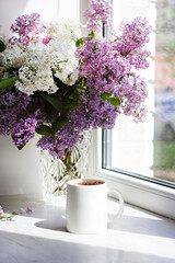 A bouquet of flowers on the window and coffee in a white cup in the rays of sunlight. A bouquet of lilacs and a cup of coffee. - 436735811