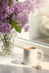 A bouquet of flowers on the window and coffee in a white cup in the rays of sunlight. A bouquet of lilacs and a cup of coffee. - 436735889