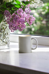 A bouquet of flowers on the window and coffee in a white cup in the rays of sunlight. A bouquet of lilacs and a cup of coffee. - 436736036