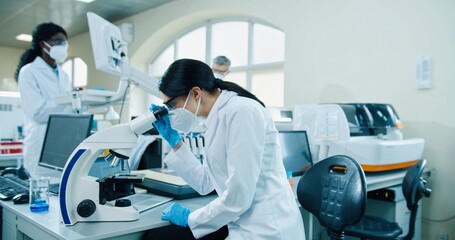 Close up of young Caucasian woman medical scientist in mask and white coat sitting at clinic laboratory using microscope looking at blood cells and looking at camera while colleague work on background