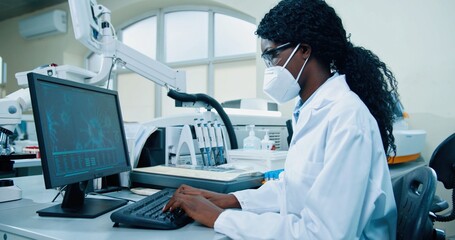 Side view of busy African American young female medical researcher sitting in lab working typing on...