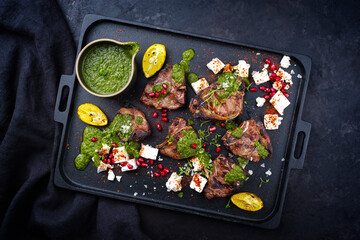 Traditional barbecue T-Bone lamb steaks with Greek feta cheese, chimichurri sauce and pomegranate fruit served as top view on a design black tray