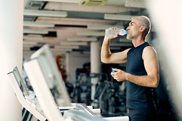 Thirsty mature athlete drinking water after running on treadmill in a gym.