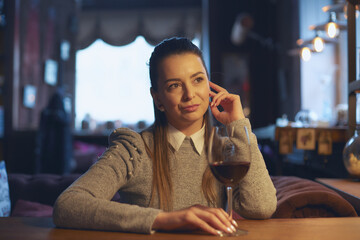 A beautiful young woman sits at a wooden table in the premises of a wine bar and drinks wine from a glass.