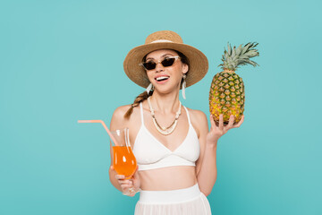 Cheerful woman in sunglasses holding pineapple and cocktail isolated on blue.