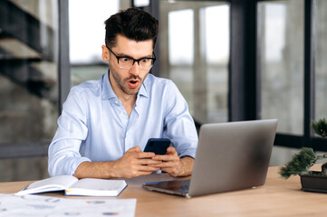 Pleasantly shocked caucasian man with, manager, broker or freelancer, sitting in the office, using mobile phone, surprised by news or message, looking at screen in amazement, tearing his mouth
