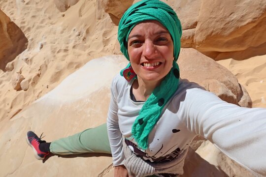 A cheerful young woman wearing a white shirt and a green head cover  with hazel eyes taking a selfie in the middle of the rocks of the white canyon in Egypt
