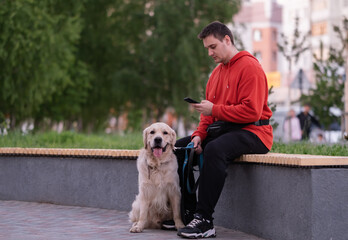 A guy in a red sweatshirt walks around the city with a dog. A young man sits on the phone on a...