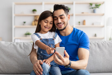 Smiling dad and daughter sitting on couch, using phone - Powered by Adobe