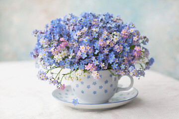 A bouquet of spring flowers forget-me-nots in a cup on the table against the background of a decorative colored wall. Postcard, blur, selective focus. - Powered by Adobe