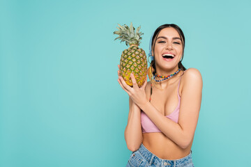 Positive woman in top holding pineapple and looking at camera isolated on blue.