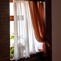 Stylish white and brown curtains hang on window with lush houseplants on sill in room decorated with red bricks in modern house