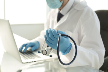 Doctor in blue gloves holds a stethoscope in one hand with the other hand is typing on a laptop in a hospital room