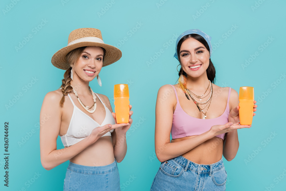 Wall mural pretty women in tops pointing at sunscreen isolated on blue.