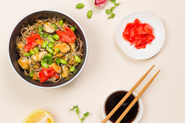 A black bowl of buckwheat soba noodles with seafood and vegetables. Flatlay on a light background with soy sauce and chopsticks. Close up