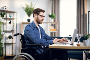 Side view of bearded young man sitting in wheelchair and typing on wireless laptop. Handsome...