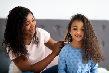 Beautiful black woman brushing her daighter's hair on bed indoors