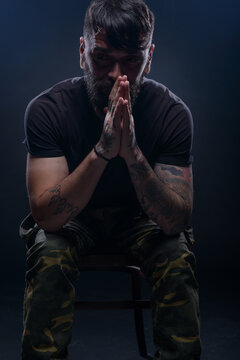 Closeup dark picture of a bearded male model in black t-shirt  sitting on a chair and praying with his tattooed hands