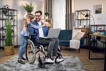 Fototapeta na wymiar Cute daughter embracing her beloved father sitting in wheelchair and using modern laptop. Handicapped man working from home to support his family.