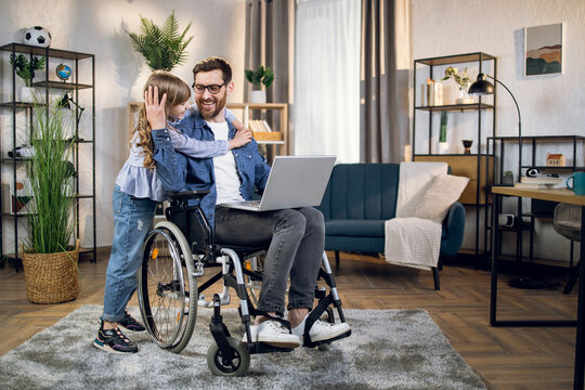 Cute Daughter Embracing Her Beloved Father Sitting In Wheelchair And Using Modern Laptop. Handicapped Man Working From Home To Support His Family.